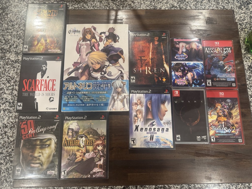 Flat-lay of Dakota's 27th birthday haul: PS2 games, Switch titles, and an Ar Tonelico art book on a wooden table